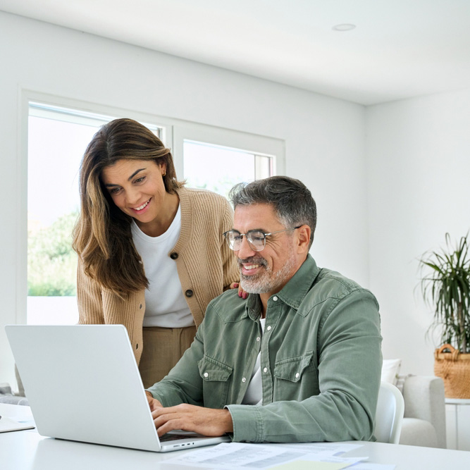 woman standing beside sitting man, both smiling while looking at laptop screen